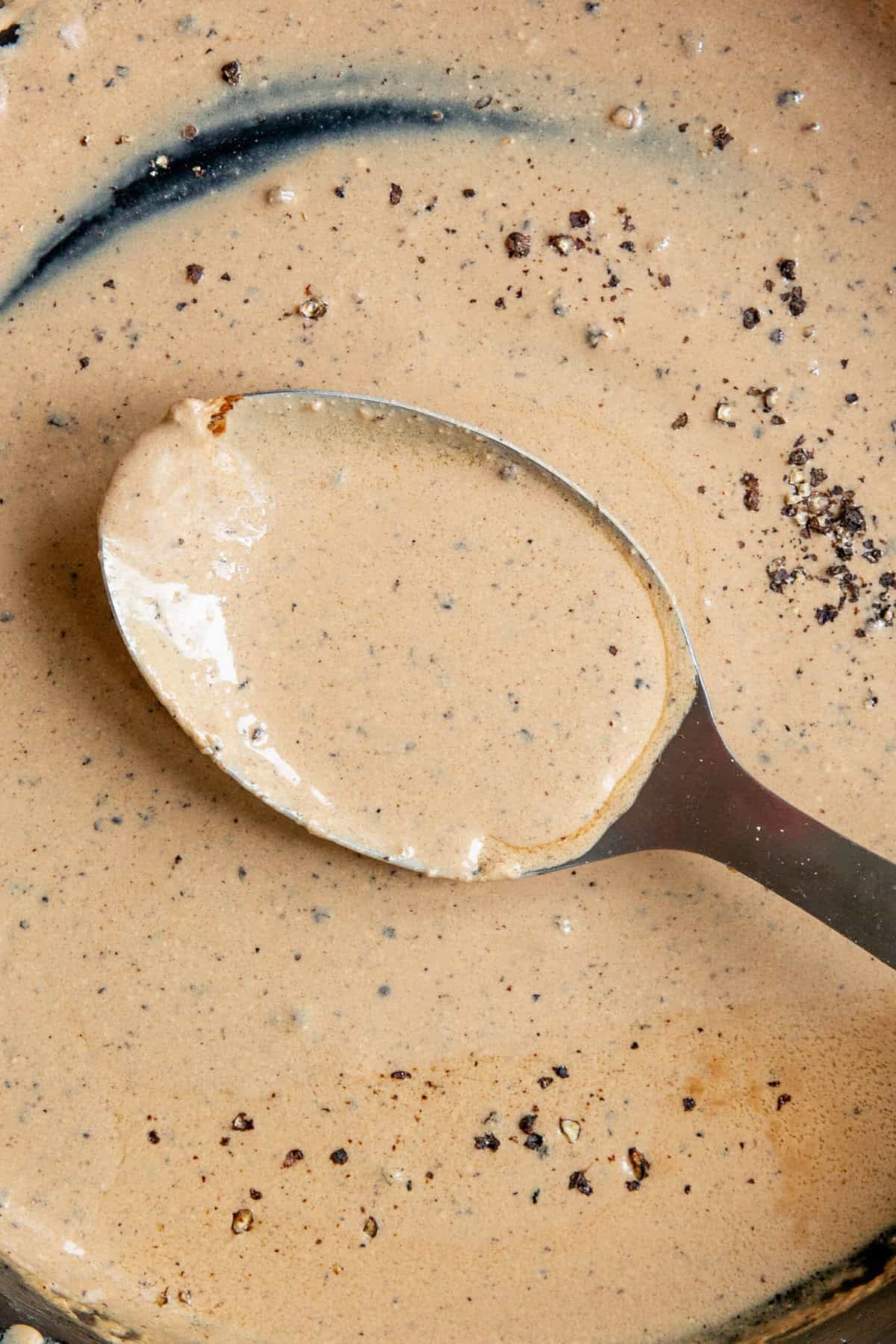 A close-up of a spoon with creamy brown peppercorn sauce for steak in a pan, with visible specks of ground black pepper scattered on the sauce’s surface.