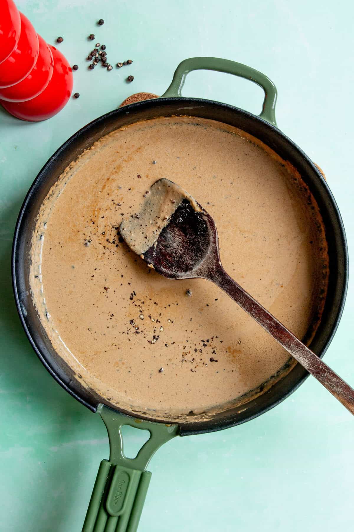 A green-handled skillet with creamy Peppercorn Sauce for Steak being stirred by a wooden spoon, surrounded by whole black peppercorns and a red pepper mill on a light green surface.
