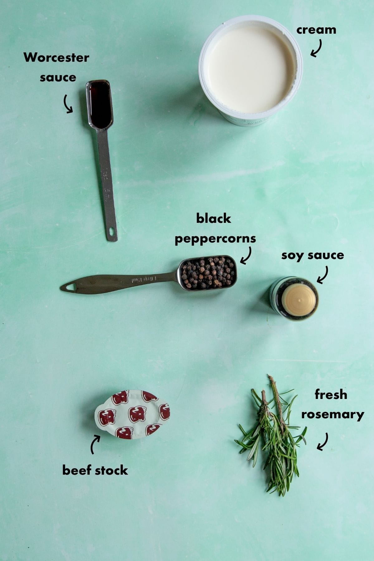 Ingredients for Peppercorn Sauce for Steak on a green surface: a spoon of Worcester sauce, a container of cream, a spoon of black peppercorns, a bottle of soy sauce, a bowl of beef stock, and sprigs of fresh rosemary.