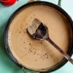 A cast iron skillet with creamy Peppercorn Sauce for Steak being stirred by a wooden spoon, topped with cracked black pepper. A red salt shaker sits nearby on a light green surface.