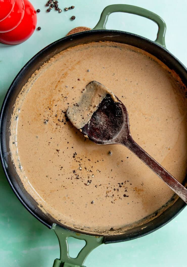 A cast iron skillet with creamy Peppercorn Sauce for Steak being stirred by a wooden spoon, topped with cracked black pepper. A red salt shaker sits nearby on a light green surface.