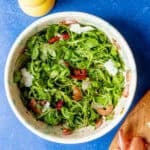 A bowl of rocket salad with shaved parmesan, sun-dried tomatoes, and sliced shallots sits on a blue surface beside a wooden cutting board with shallot pieces and a yellow pepper grinder.