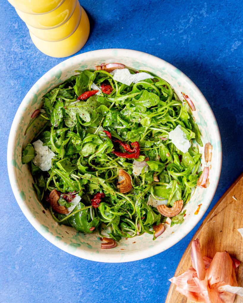 A bowl of rocket salad with shaved parmesan, sun-dried tomatoes, and sliced shallots sits on a blue surface beside a wooden cutting board with shallot pieces and a yellow pepper grinder.