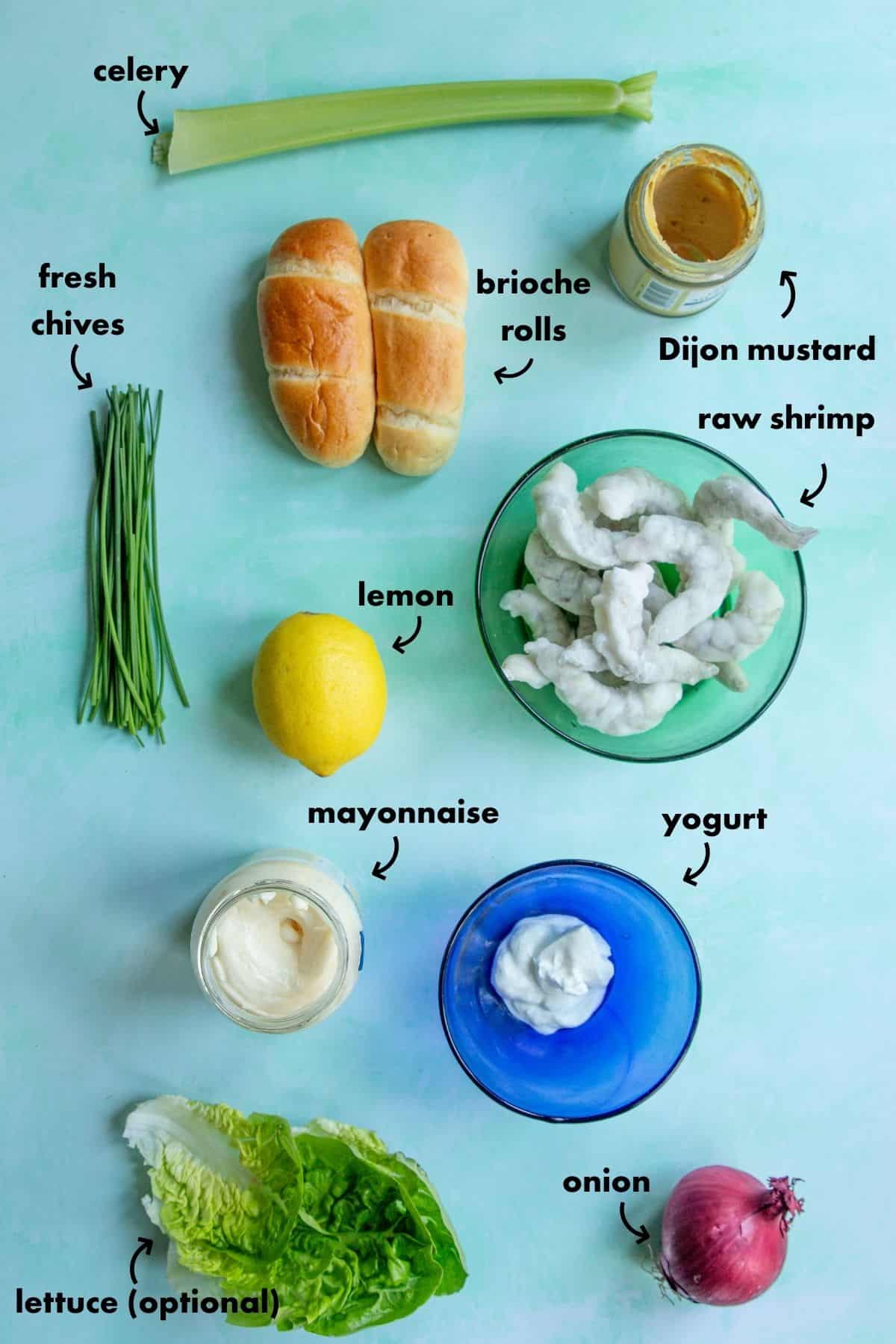 A top-down view of ingredients for a shrimp roll: celery, fresh chives, brioche rolls, Dijon mustard, raw shrimp, lemon, mayonnaise, yogurt, red onion, and lettuce—all labeled on a light blue background.
