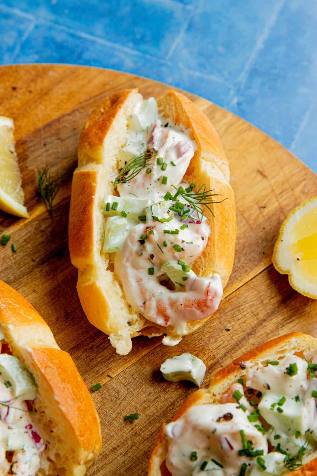 A close-up of a shrimp sandwich on a wooden board, filled with shrimp, chopped celery, herbs, and a creamy dressing, served with lemon wedges on the side.