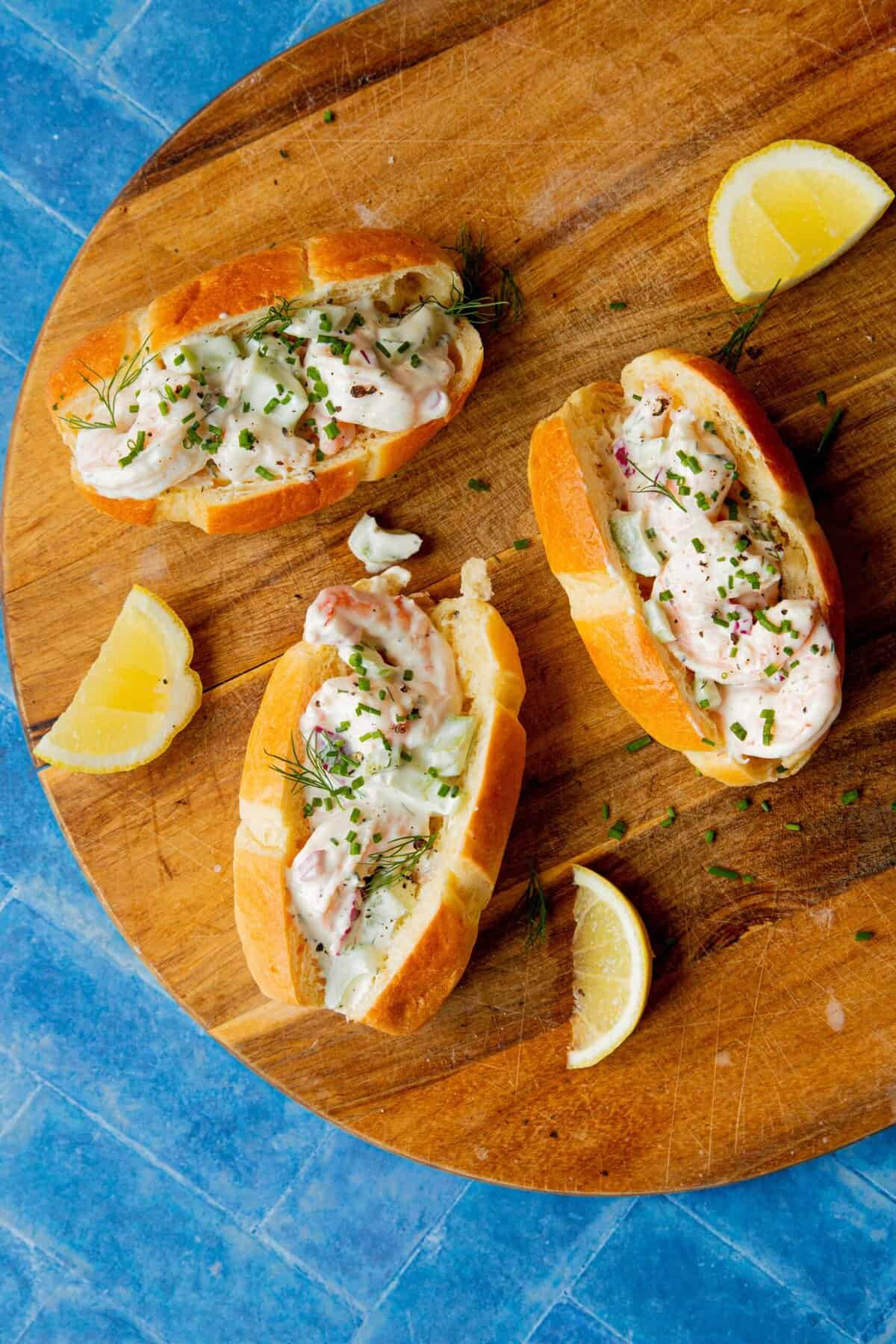 Three shrimp rolls topped with creamy sauce and herbs are arranged on a round wooden board with lemon wedges, set against a blue tiled background.