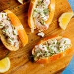 Three lobster rolls filled with creamy lobster salad and garnished with herbs on a round wooden board, served alongside lemon wedges. The background is a blue textured surface.