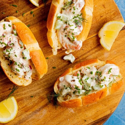 Three lobster rolls filled with creamy lobster salad and garnished with herbs on a round wooden board, served alongside lemon wedges. The background is a blue textured surface.