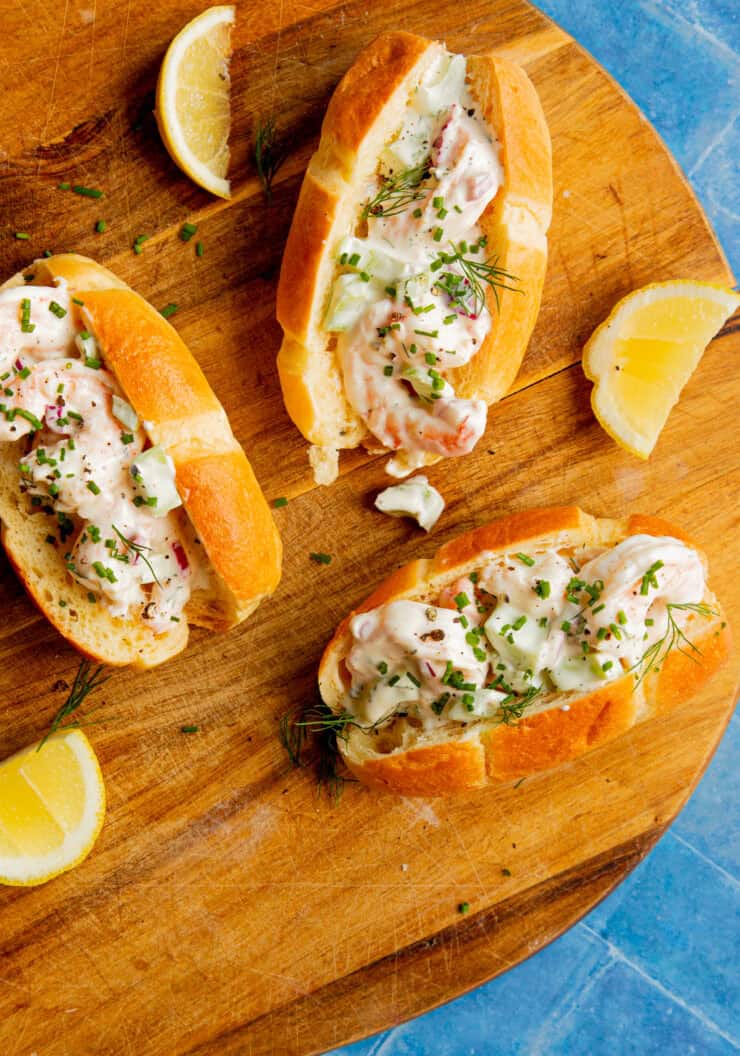 Three lobster rolls filled with creamy lobster salad and garnished with herbs on a round wooden board, served alongside lemon wedges. The background is a blue textured surface.