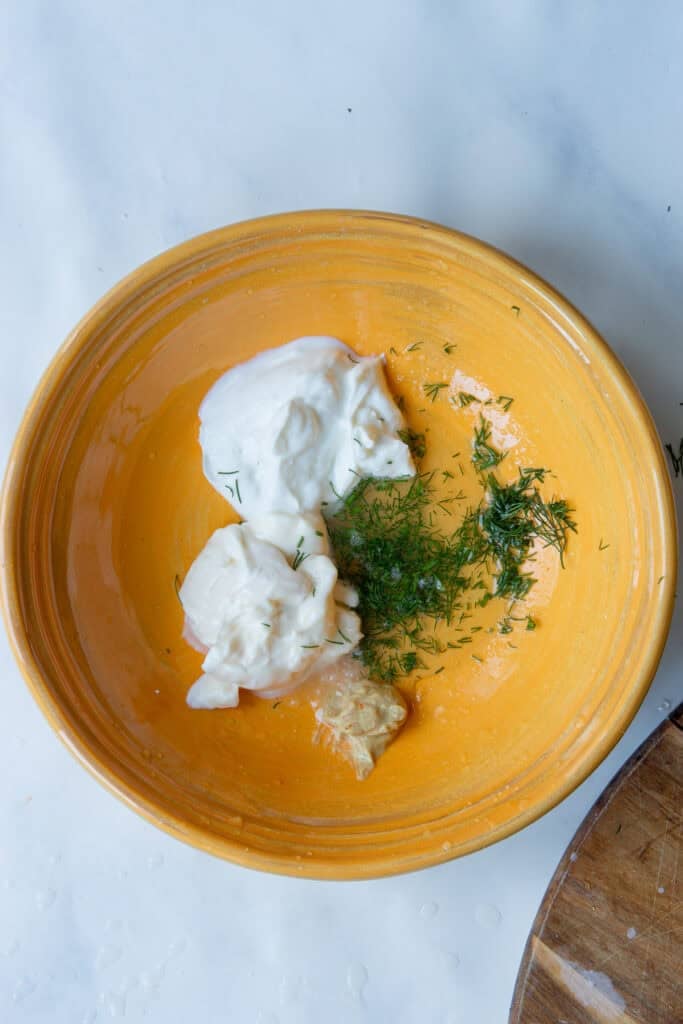 A yellow bowl containing plain yogurt, mayonnaise, minced garlic, and chopped fresh dill sits on a white surface with part of a wooden cutting board visible at the edge.