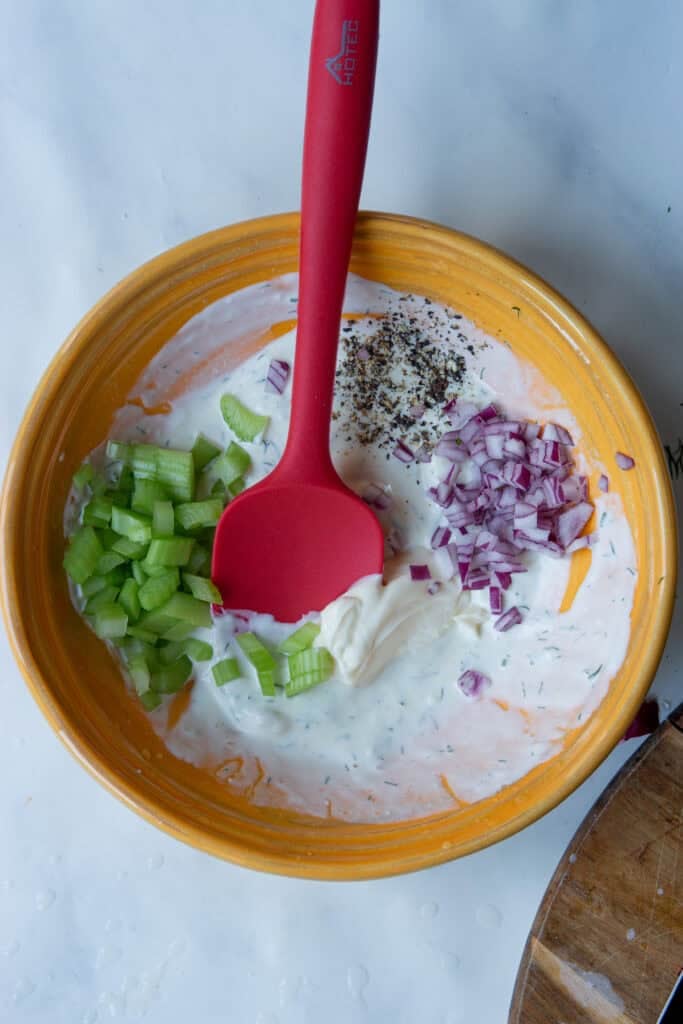An orange bowl holds yogurt, mayonnaise, chopped celery, diced red onion, and ground black pepper. A red spatula rests in the bowl, ready to mix the ingredients.