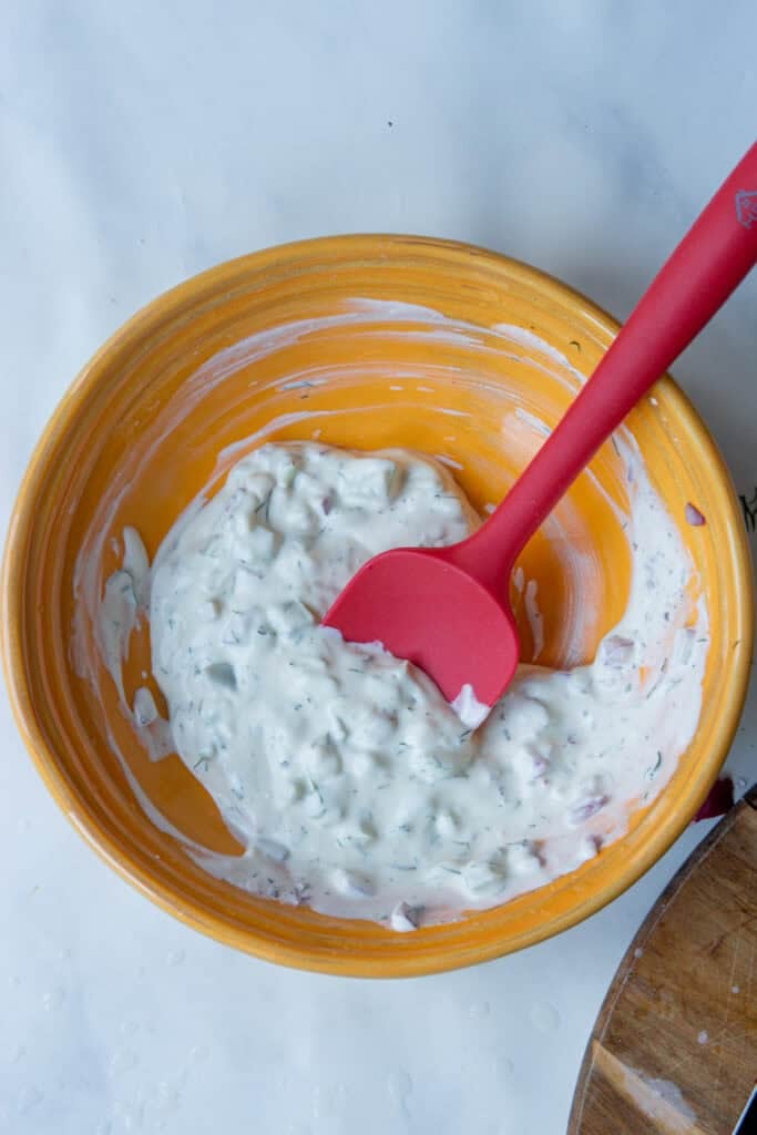 An orange bowl filled with creamy tzatziki sauce, mixed with chopped herbs, cucumber, sits on a white surface. A red spatula rests inside the bowl, partially covered with the sauce.