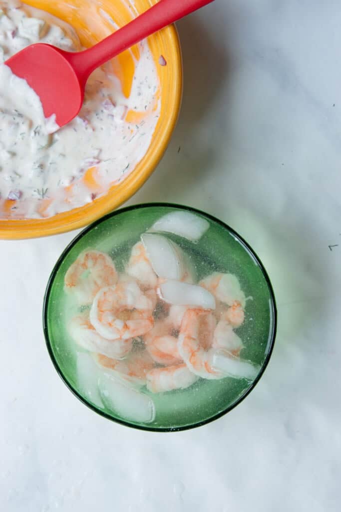 A green bowl filled with cooked shrimp and ice cubes sits beside a yellow bowl containing a creamy herb sauce being mixed with a red spatula on a white surface.
