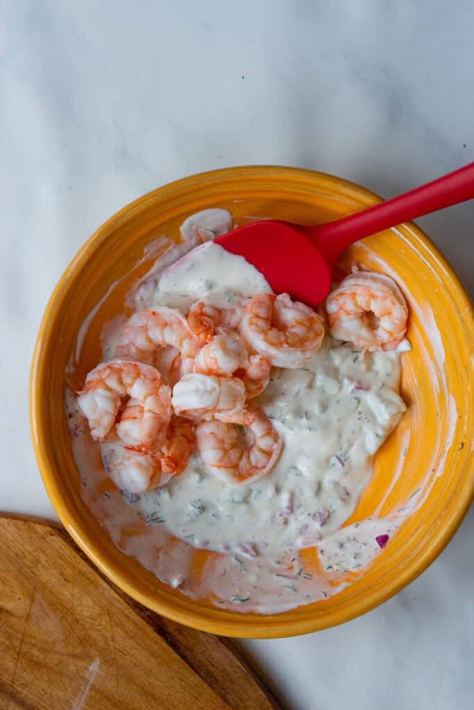 An orange bowl with shrimp in a creamy white sauce, mixed with herbs and diced red onions sits on a light surface. A red spatula rests in the bowl. Part of a wooden board is visible in the corner.
