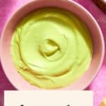 A bowl of creamy Avocado Mayo sits on a pink surface, surrounded by half an avocado, a lemon wedge, and a jar. Text reads Avocado Mayo At Home.