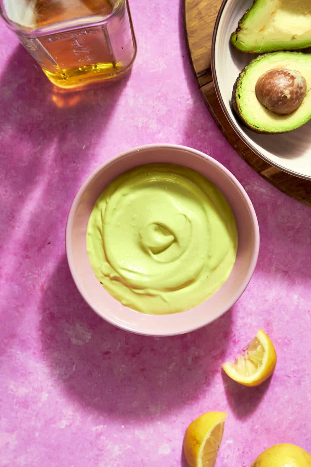 A bowl of creamy avocado sauce sits on a pink surface, surrounded by roasted corn, lemon wedges, a bottle, and a plate with halved avocados.