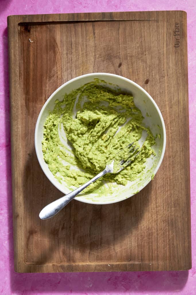 A white bowl containing mashed avocado with roasted corn and a fork rests on a wooden cutting board, set against a bright pink background.