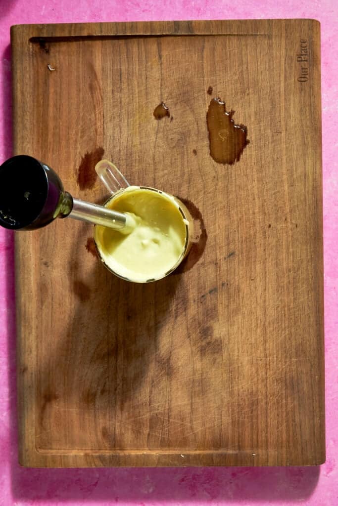 A glass measuring cup with creamy roasted corn sauce being blended by an immersion blender sits on a wooden cutting board, which has a few liquid stains, against a pink background.