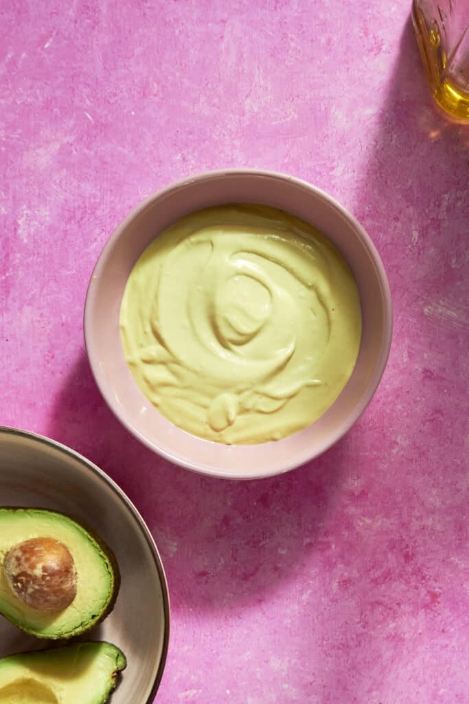 A pink bowl filled with creamy avocado sauce sits on a pink textured surface. Next to it, a plate holds roasted corn and a halved avocado, while part of a bottle, likely oil, is visible in the corner.