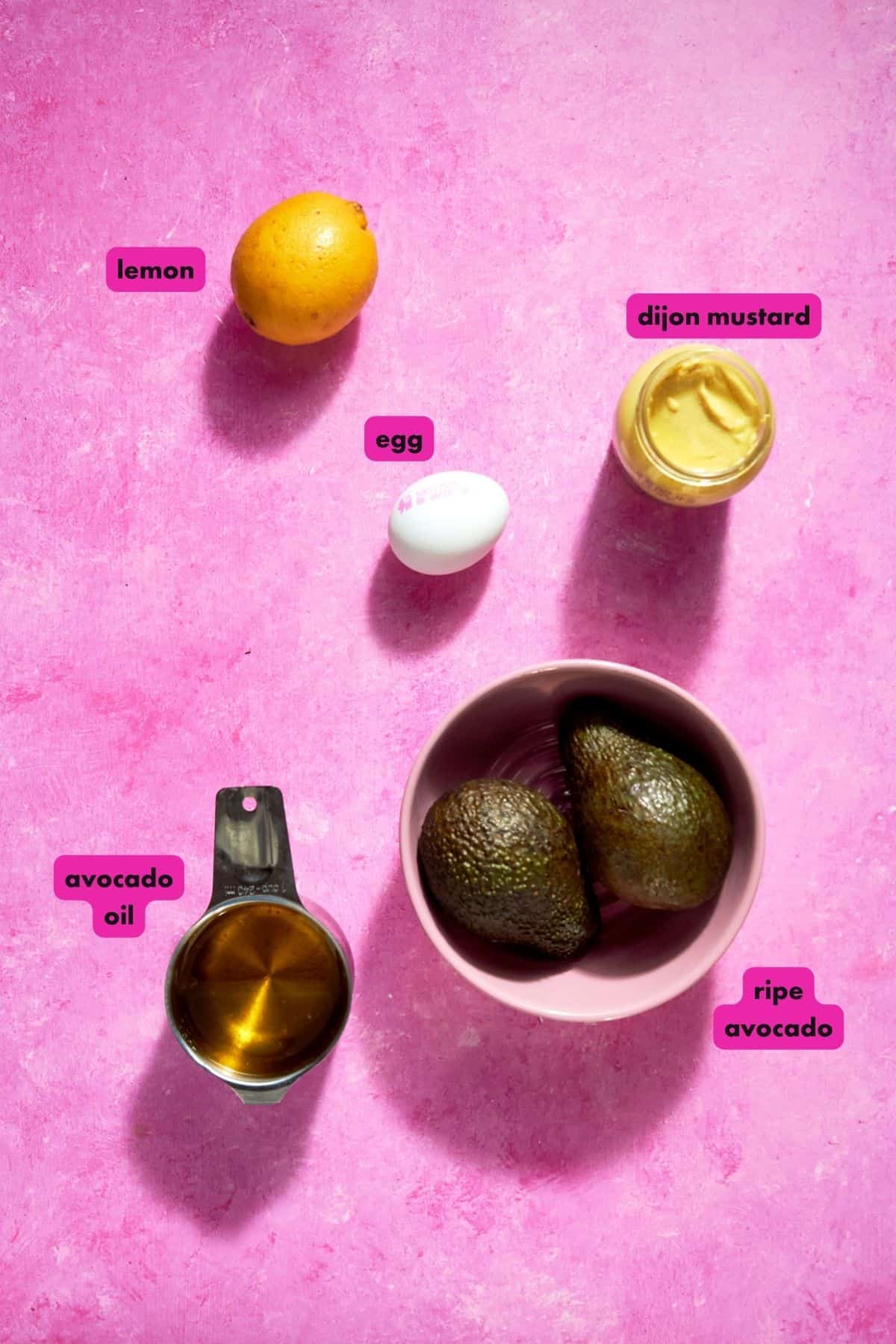 A flat lay of ingredients on a pink background: a lemon, a jar of Dijon mustard, an egg, roasted corn, a bowl with two ripe avocados, and a measuring cup with avocado oil. Each item is labeled with pink tags.
