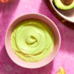 A pink bowl filled with creamy, light green avocado dip sits on a pink surface next to roasted corn, a halved avocado, a lemon wedge, and a bottle of oil.