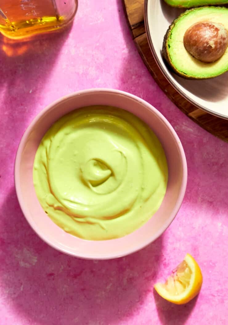 A pink bowl filled with creamy, light green avocado dip sits on a pink surface next to roasted corn, a halved avocado, a lemon wedge, and a bottle of oil.