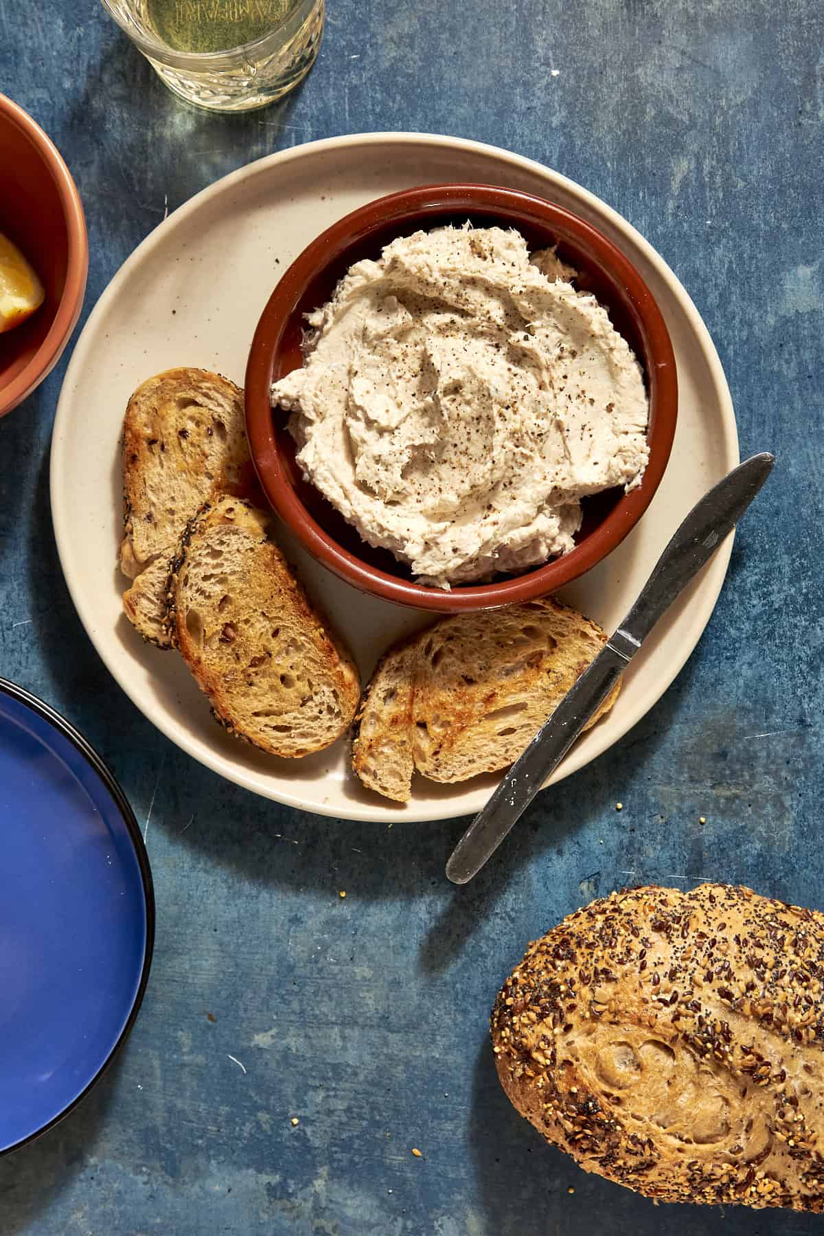 A bowl of whipped spread, topped with black pepper and hints of roasted corn, sits on a plate with slices of toasted bread. A knife rests on the plate, with a seeded loaf and glass of water nearby on a blue surface.