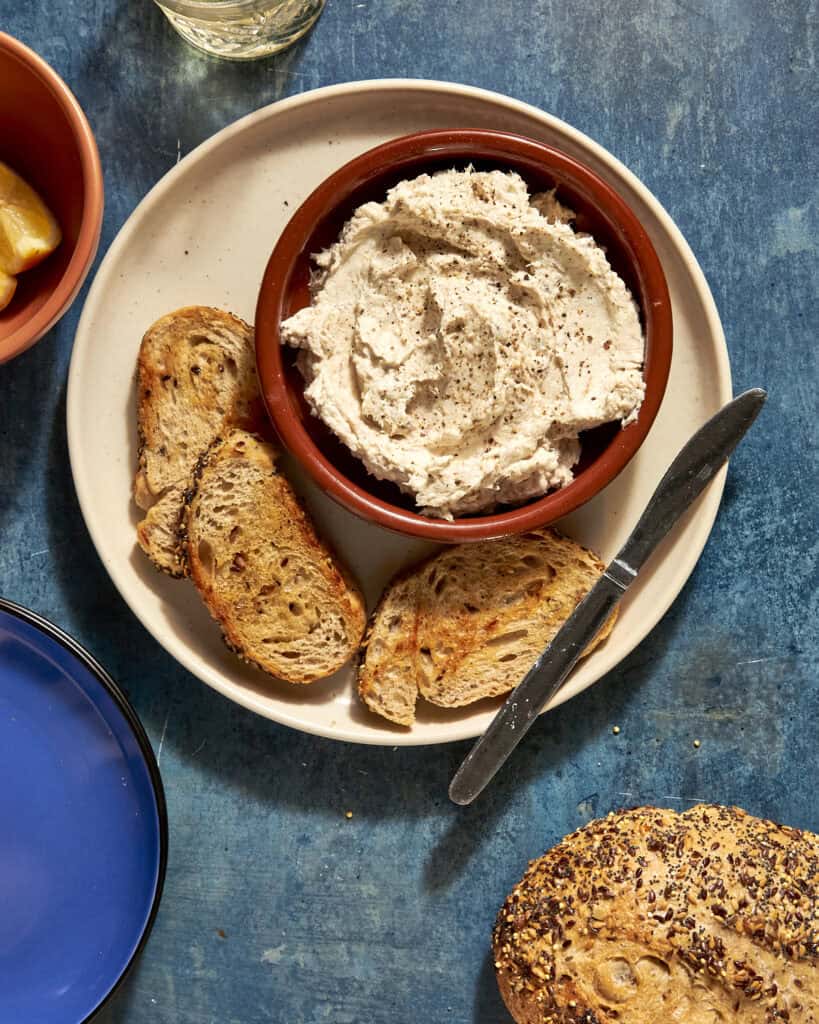 A bowl of creamy roasted corn spread with black pepper on top sits on a white plate with three slices of seeded bread and a knife, all placed on a blue tabletop. A partially visible loaf and bowls are nearby.