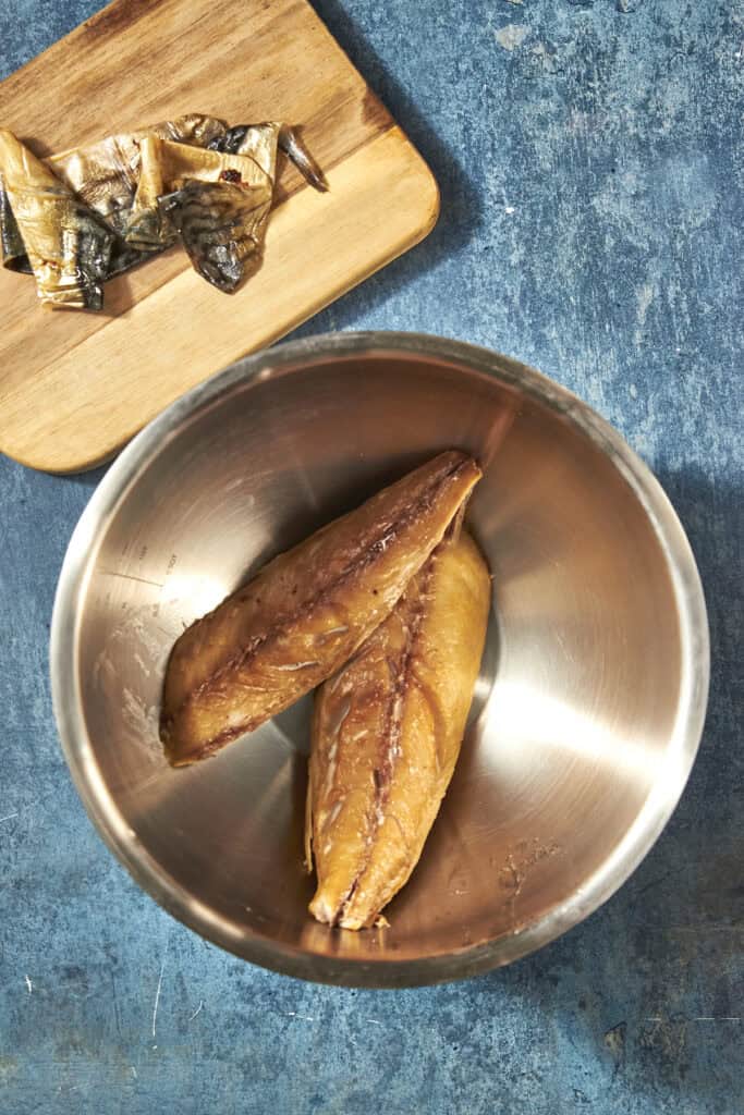 Two pieces of smoked fish in a metal bowl sit on a blue textured surface; above the bowl, a wooden cutting board holds smaller pieces of the fish alongside golden roasted corn.