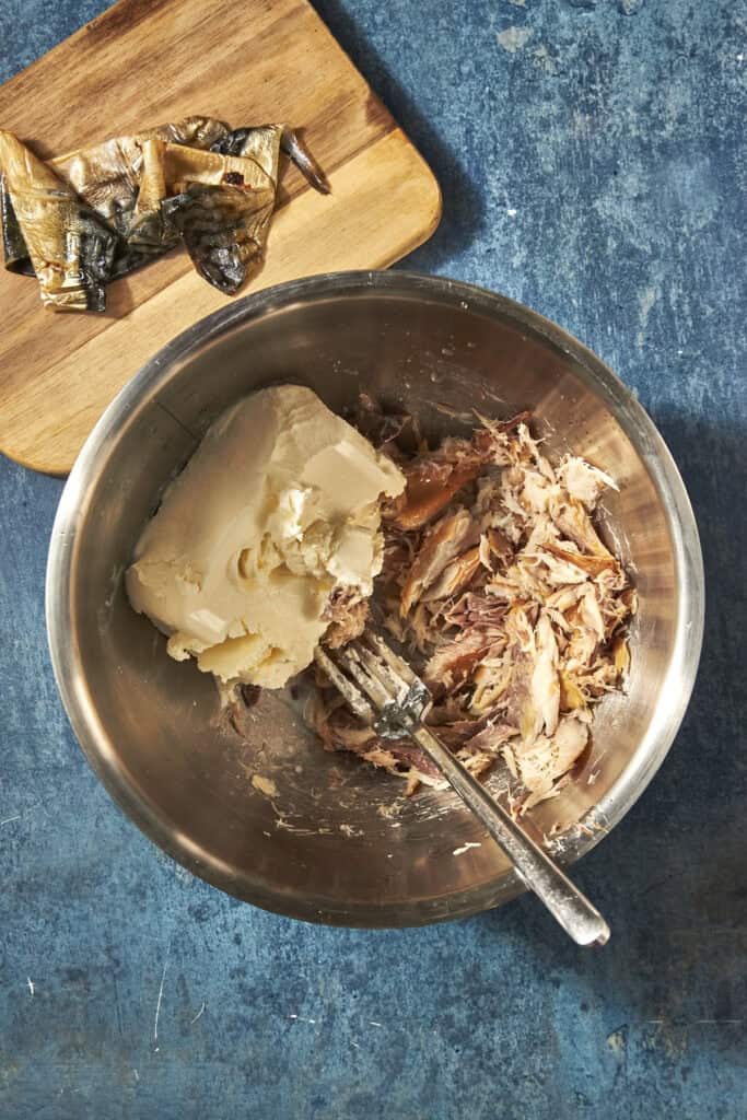 A metal bowl containing chunks of butter, flaked fish, and hints of roasted corn, with a fork resting inside; in the background, a wooden board holds pieces of smoked fish skin, all set on a blue textured surface.