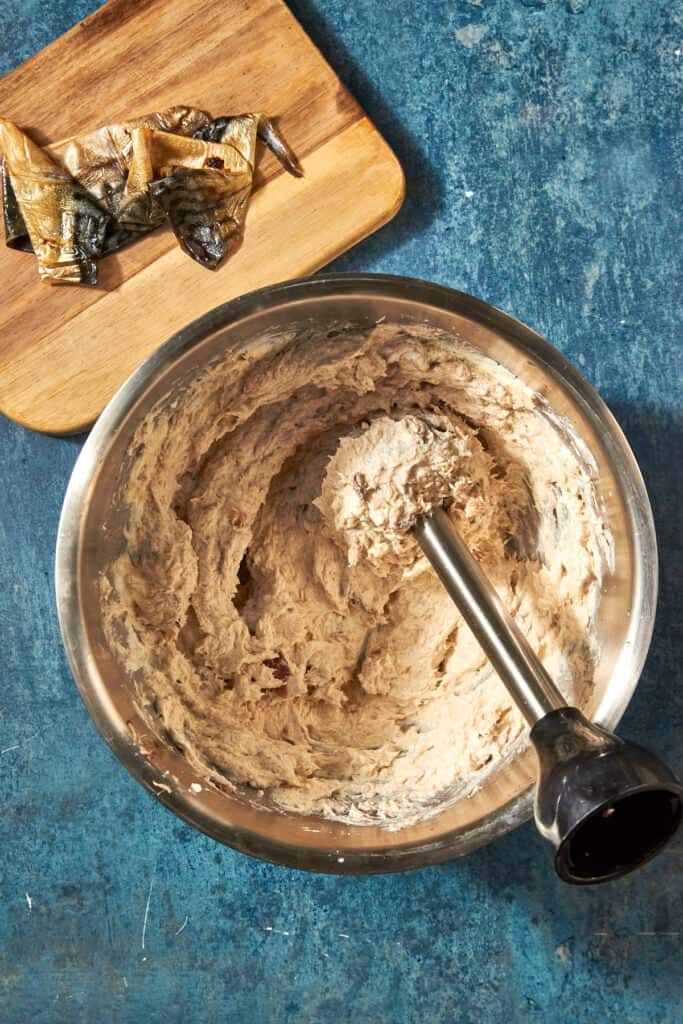 A mixing bowl filled with a creamy, beige mixture and roasted corn is being blended with an immersion blender. In the background, pieces of fish skin rest on a wooden cutting board, all set on a blue textured surface.