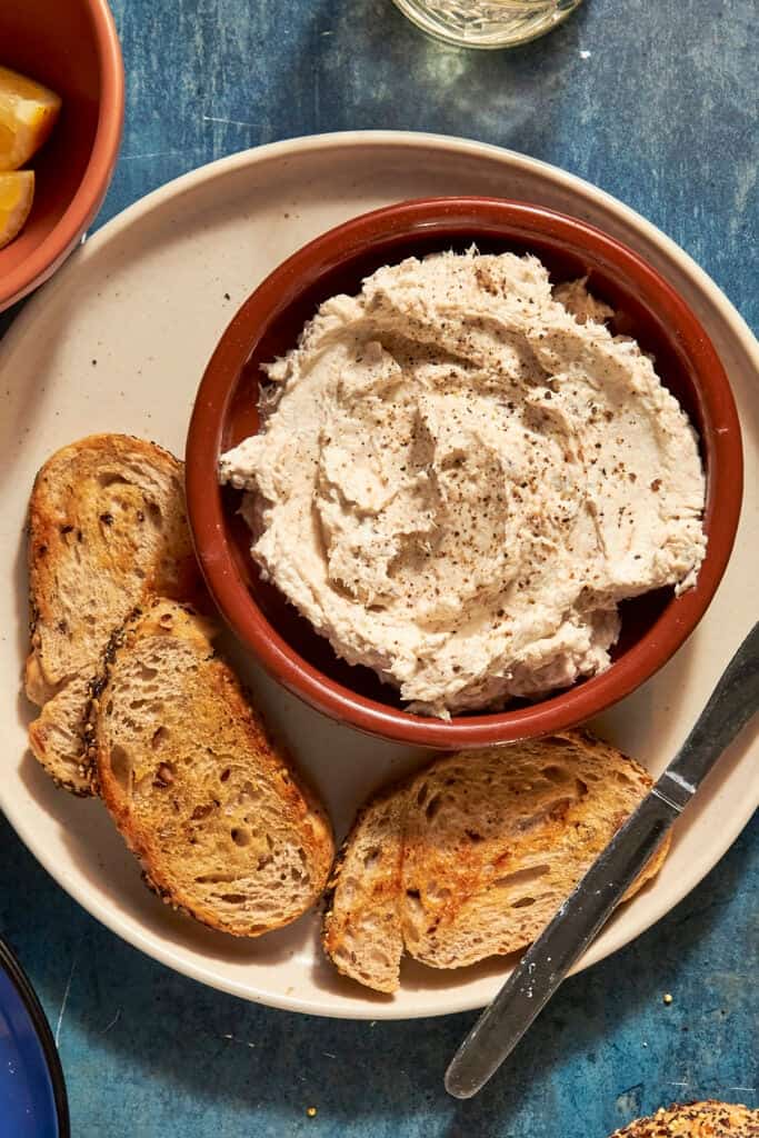 A bowl of creamy dip with roasted corn and a sprinkle of black pepper sits on a plate with slices of toasted bread and a butter knife, set on a blue surface.