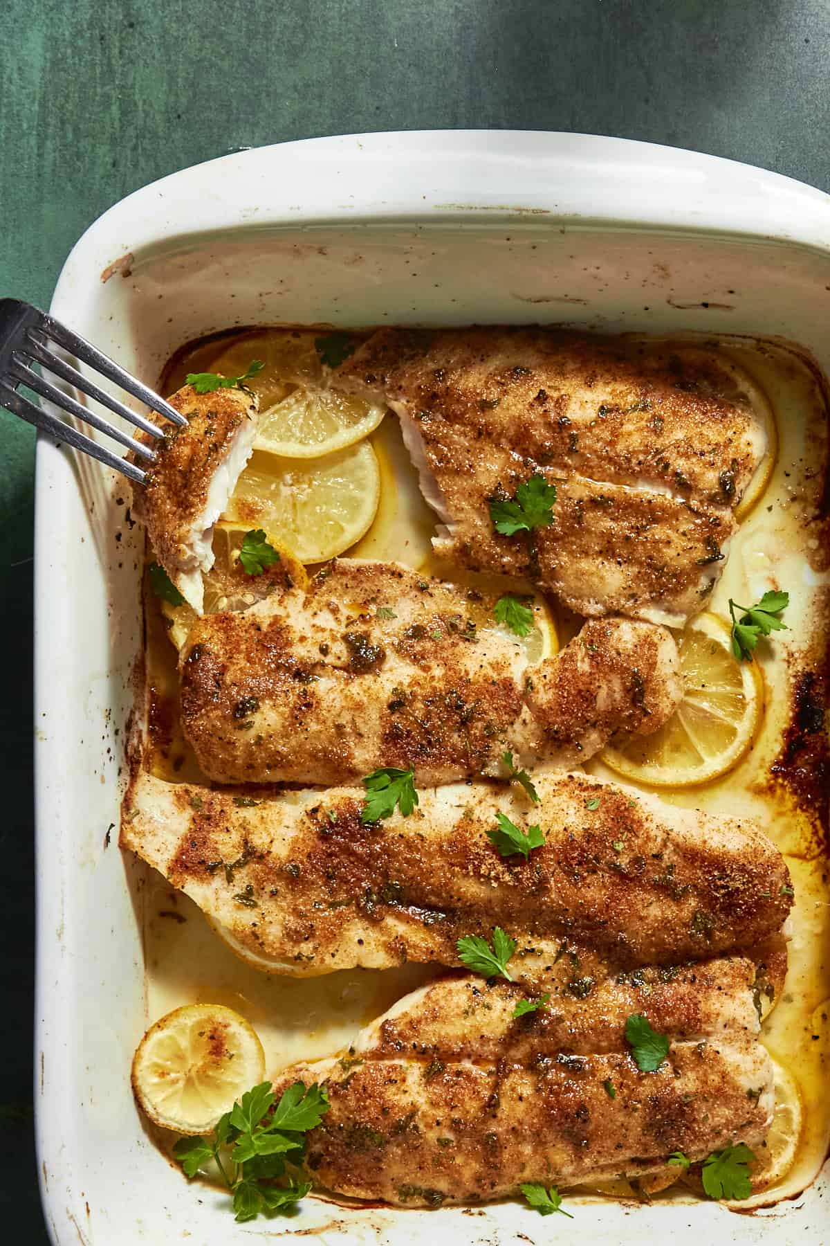 Baked seasoned fish fillets in a white dish, garnished with fresh parsley, lemon slices, and roasted corn. A fork is flaking one fillet. The dish sits on a green surface.
