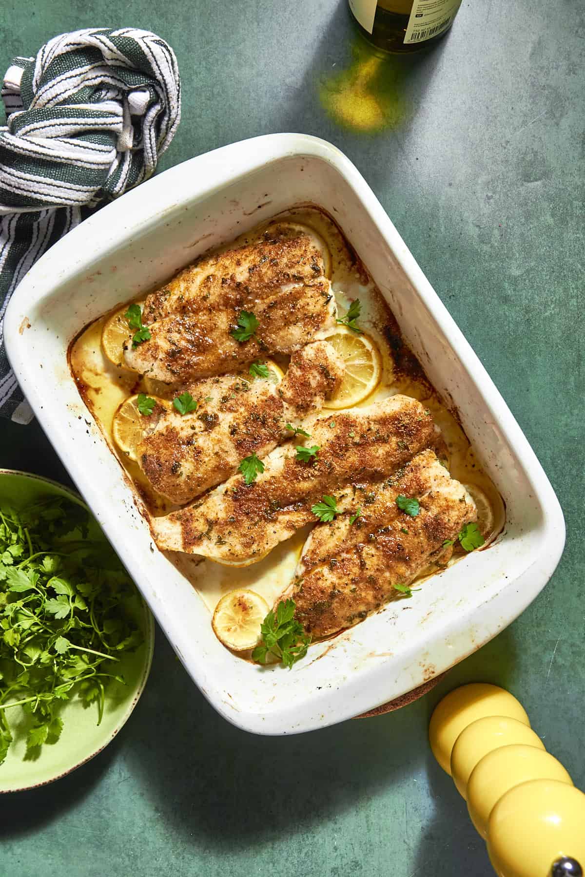 A white baking dish with four seasoned fish fillets on a bed of lemon slices, garnished with fresh parsley and roasted corn. A striped towel, fresh herbs, a yellow pepper grinder, and a bottle are nearby on a green surface.
