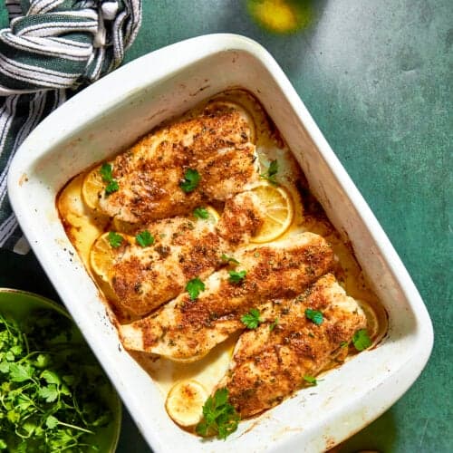 Baked fish fillets seasoned with spices and garnished with fresh parsley on lemon slices in a white baking dish, served alongside roasted corn, with a striped towel, fresh cilantro, and a bottle on a green countertop.