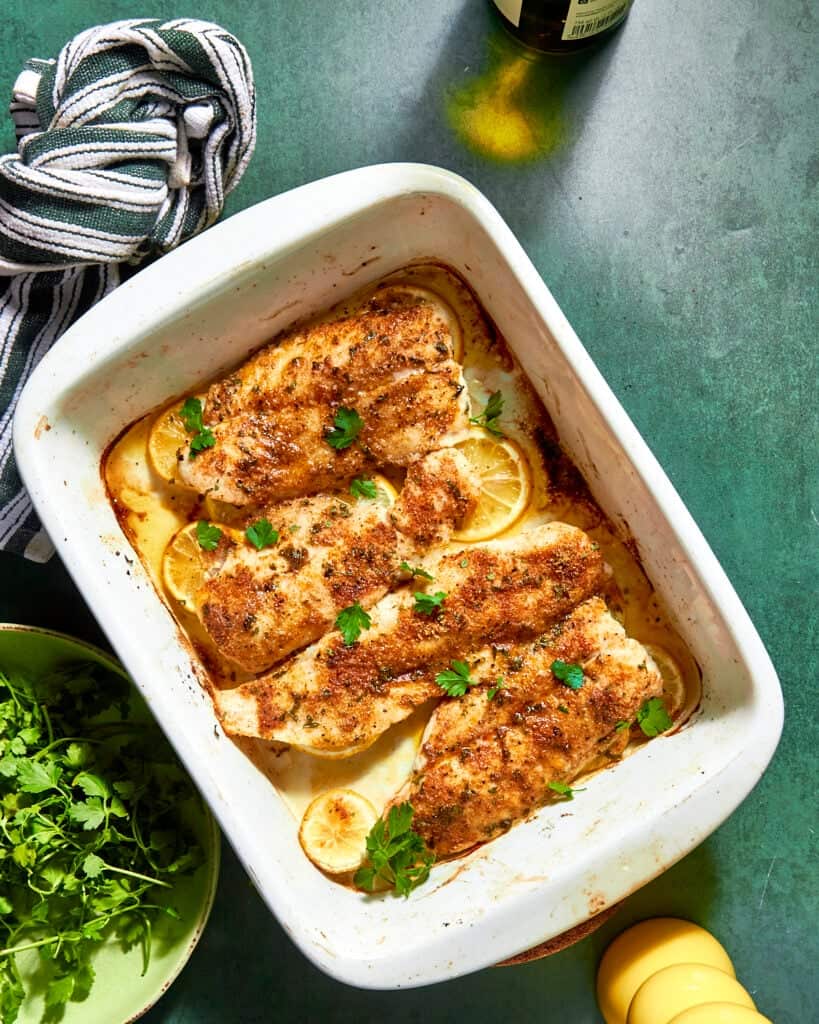 Baked fish fillets seasoned with spices and garnished with fresh parsley on lemon slices in a white baking dish, served alongside roasted corn, with a striped towel, fresh cilantro, and a bottle on a green countertop.