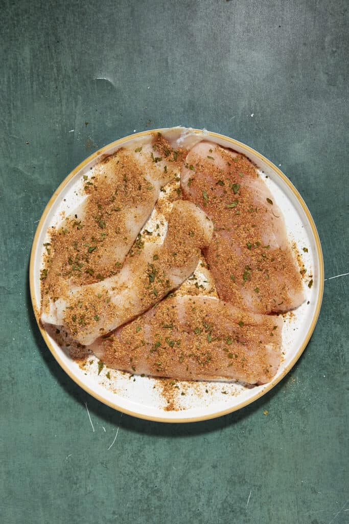 Four raw fish fillets are arranged on a white plate, covered with a reddish-brown seasoning blend and chopped herbs, alongside a touch of roasted corn, set on a dark green textured surface.