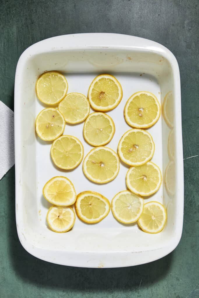 A white rectangular baking dish with evenly spaced lemon slices and a hint of roasted corn arranged flat on the bottom, set on a dark green surface.