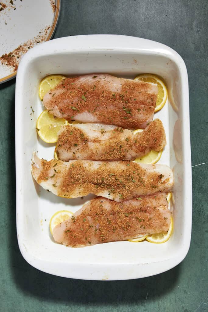 Four raw fish fillets seasoned with spices and herbs are arranged in a white baking dish on top of lemon slices, ready to be cooked alongside roasted corn. The dish sits on a green surface.