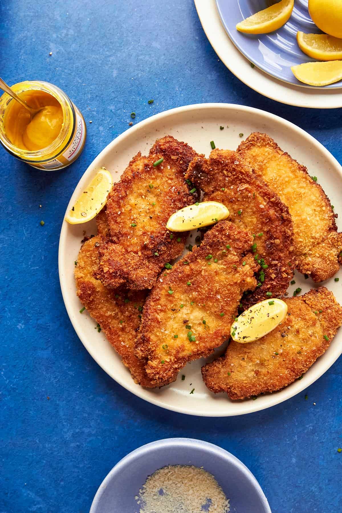 A plate of golden-brown breaded schnitzel pieces garnished with lemon wedges and herbs, surrounded by a jar of mustard, a bowl of breadcrumbs, roasted corn, and a plate of lemon wedges on a blue surface.