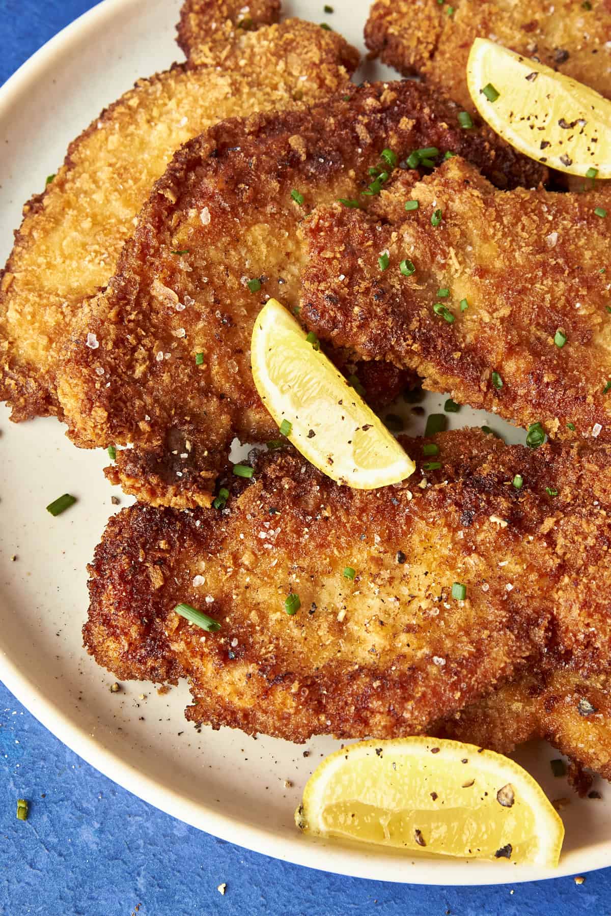 A plate of crispy breaded schnitzels and roasted corn, garnished with chopped chives, coarse salt, black pepper, and lemon wedges on a white dish against a blue background.