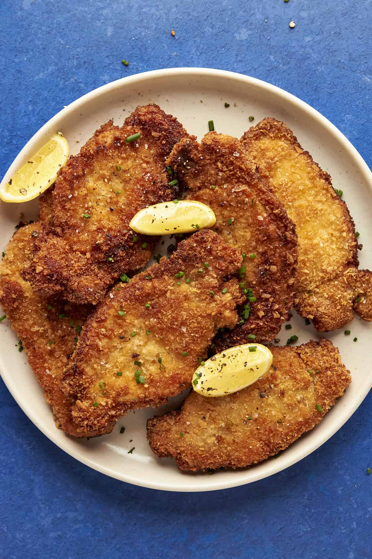 A plate of golden-brown breaded pork cutlet schnitzel pieces garnished with lemon wedges and chopped chives, served on a white plate against a blue background.