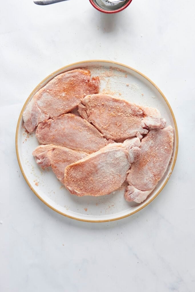 Five raw, seasoned pork chops are arranged on a round, white plate with a beige rim, placed on a white marble surface. A small portion of roasted corn in a spoon and bowl is visible at the top of the image.