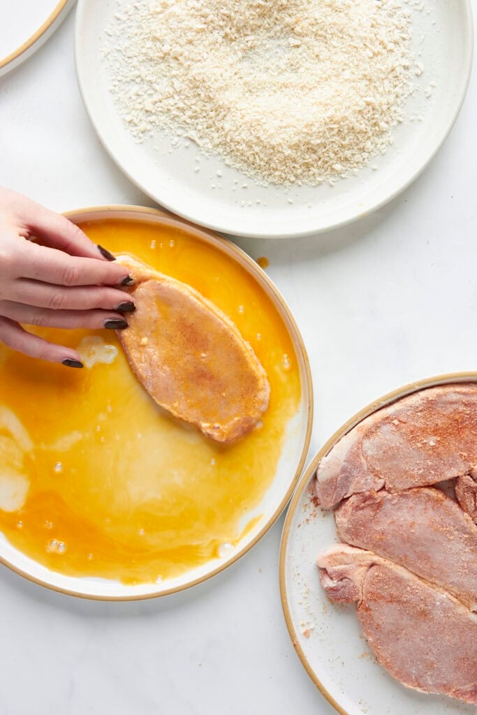 A hand dips a seasoned raw pork chop into a bowl of beaten eggs, with a plate of roasted corn bread crumbs above and more seasoned pork chops beside it, all on a white surface.