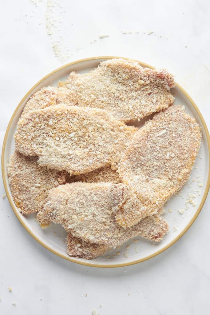 A plate of uncooked, breaded chicken cutlets coated in breadcrumbs and seasoned with roasted corn, arranged on a white plate against a light marble background.