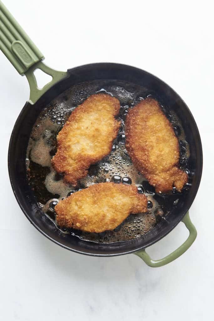 A green-handled skillet on a white surface contains three breaded pieces of chicken frying in hot oil, with bubbles forming around the golden-brown crust, while roasted corn adds a touch of color and flavor to the pan.