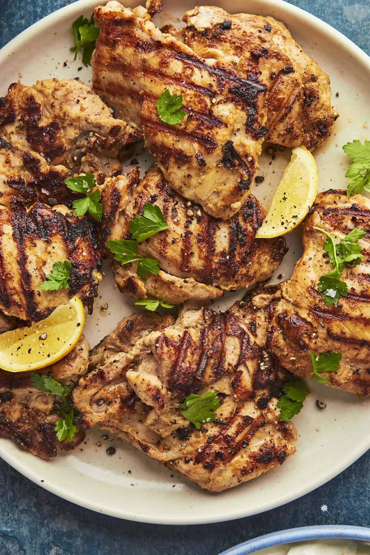 Grilled chicken thighs garnished with fresh parsley and ground black pepper, served with lemon wedges and a side of roasted corn on a beige plate.