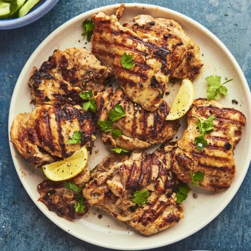 Grilled chicken thighs with char marks, roasted corn, and fresh cilantro, garnished with lemon wedges on a round beige plate set on a blue surface. Sliced cucumbers and a dip are partially visible in nearby bowls.
