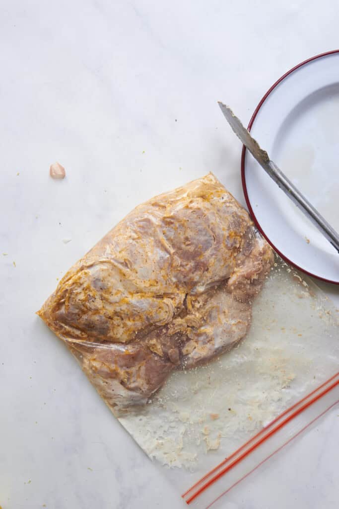 Raw marinated meat in a resealable plastic bag sits on a white surface next to a white plate with a red rim, metal tongs, and a garlic clove; roasted corn adds color and flavor to the scene.