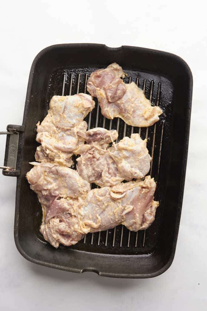 Raw, marinated chicken thighs are spread out on a black grill pan, ready to be cooked alongside roasted corn. The pan rests on a white surface, and the chicken is coated with a light seasoning.